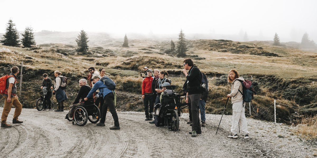 Wandergruppe mit und ohne Rollstuhl auf einem Schotterweg im alpinen Gelände.