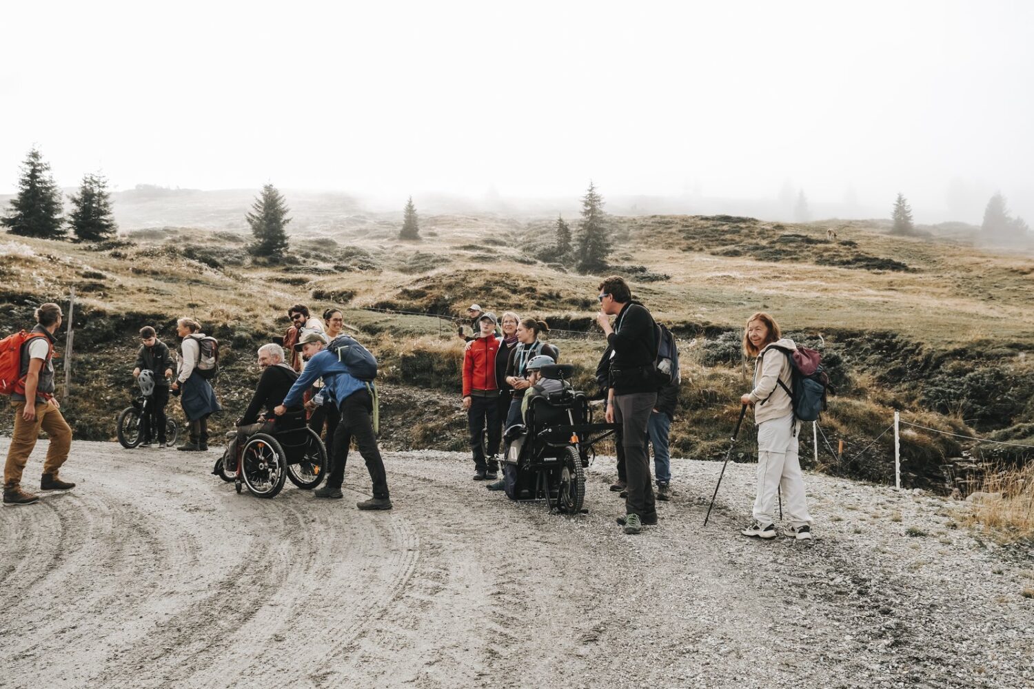Wandergruppe mit und ohne Rollstuhl auf einem Schotterweg im alpinen Gelände; zwei Personen sind im Rollstuhl unterwegs und werden von Begleitpersonen unterstützt.