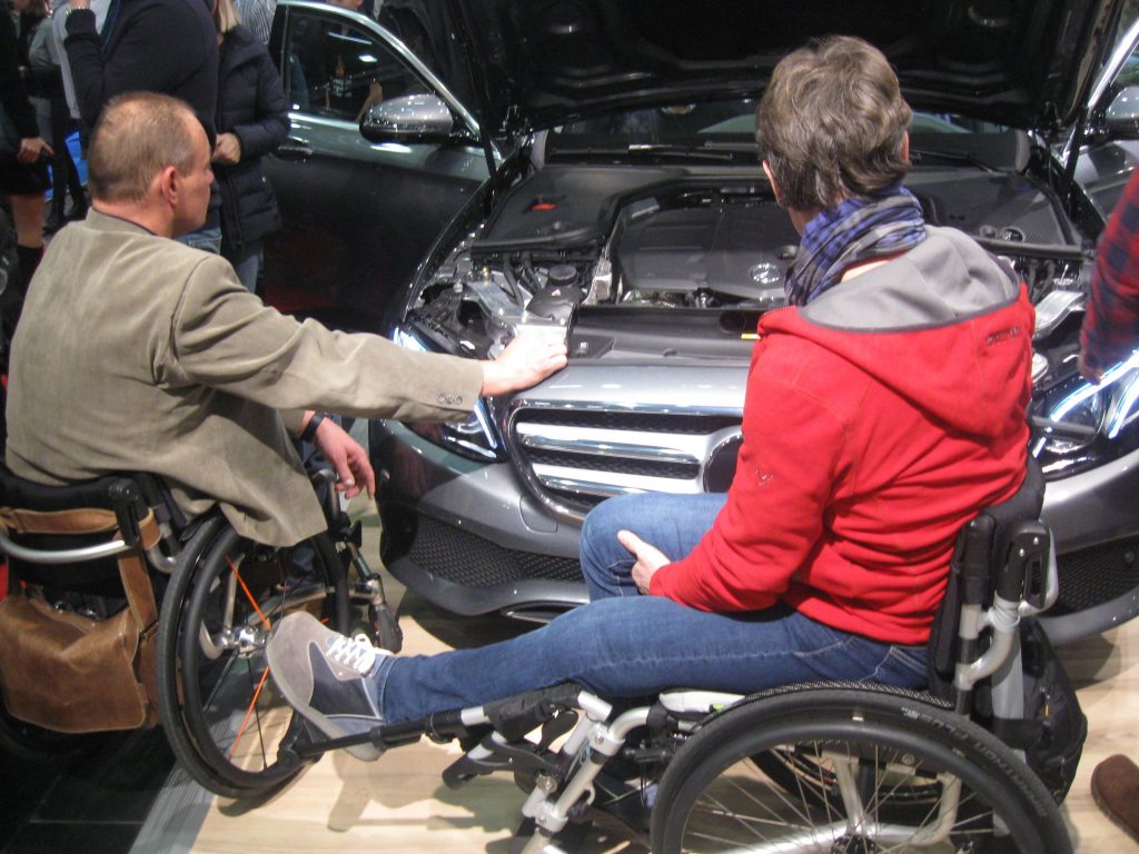 Zwei Rollstuhlfahrer betrachten die geöffnete Motorhaube eines Mercedes